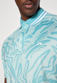 Polo de cor azul-clara com padrões ondulados abstratos, apresentando uma pala com dois botões, colarinho listrado branco em contraste e detalhe do logótipo.