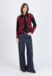 Cárdigan negro con patrón floral en rojo y rosa, llevado sobre una camiseta negra y combinado con pantalones de mezclilla oscuros de pierna ancha. Detalles de costura sutiles visibles.