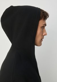 Hanorac negru cu fermoar, confecționat dintr-un material texturat, cu glugă reglabilă și croială slim, prezentat în profil lateral pe un model.