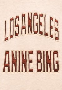 Το κείμενο «LOS ANGELES» σε καμπύλη πάνω από το «ANINE BING» με μεγάλα, καφέ, έντονα γράμματα σε μπεζ υφασμάτινο φόντο.