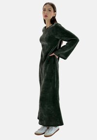 Langes, dunkelgrünes Kleid mit lockerem Schnitt, weiten Ärmeln und strukturiertem Stoff, kombiniert mit weißen Turnschuhen für einen lässigen Look.