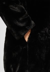Main partiellement insérée dans la poche latérale d'un manteau noir en fausse fourrure pelucheuse.