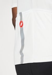 T-shirt de sport blanc avec un tissu texturé, une rayure grise courbée sur le côté et un logo circulaire rouge avec un design de scorpion.