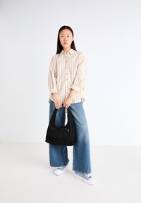 Stribet beige skjorte med knapper, vidde blå jeans og en sort skuldertaske. Modellen bærer hvide og lyseblå sneakers. Enkel baggrund.
