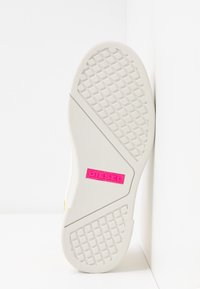 Semelle de chaussure blanche avec un motif en losanges et un logo rectangulaire rose Diesel au centre, positionnée verticalement contre un coin blanc.