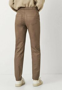 Braune Slim-Fit-Hosen mit einem elastischen Bund, zwei Gesäßtaschen und einer glatten Textur, kombiniert mit weißen Sneakers mit goldenen Akzenten.