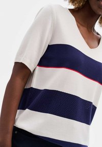 Korte mouwen trui in crème met marineblauwe en rode horizontale strepen, met een V-hals, gebreide textuur en ademende mesh-details.