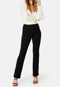Frau in schwarzen, hoch taillierten, geraden Hosen, weißer Longsleeve-Bluse mit puffigen Ärmeln und schwarzen, offenen High Heels, posiert mit der Hand nahe dem Gesicht.