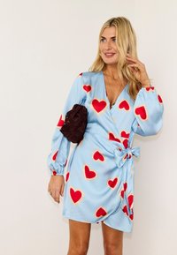 Ξανθιά γυναίκα που φοράει ένα ανοιχτό γαλάζιο φόρεμα τύπου wrap με κόκκινα σχέδια καρδιάς, κρατώντας μια μικρή σκούρα καφέ τσάντα κάτω από το μπράτσο της, χαμογελώντας, σε λευκό φόντο.