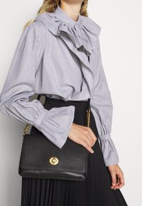 Sac à main en cuir noir avec une finition texturée, quincaillerie dorée et une bandoulière en chaîne. Le sac présente un rabat avant avec un fermoir distinctif.