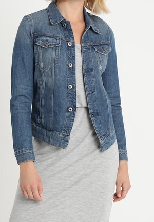 Kvinde iført en blå denimjakke med knapper foran og brystlommer over en lys grå kjole, vist fra skuldre til hofter.