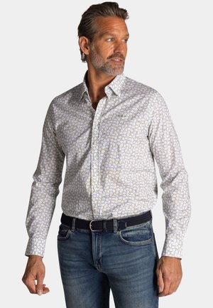 Man wearing white long-sleeve button-up shirt with small floral pattern and blue jeans, looking to his right against a plain background.