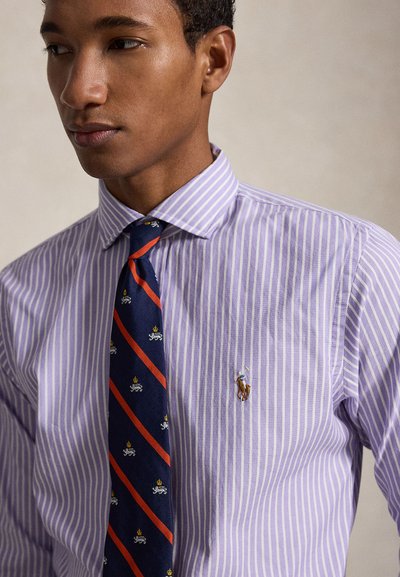 Νέος άνδρας που φοράει πουκάμισο με μοβ και λευκές ρίγες και μπλε σκούρη γραβάτα με κόκκινες ρίγες και μικρά μοτίβα λιονταριού και κορώνας.