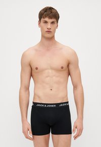 Schwarze Boxer-Shorts mit einem elastischen Markenbund in Weiß, präsentiert an einem Modell mit durchtrainiertem Körper, vor einem schlichten Hintergrund.