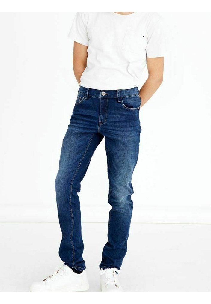 Personne portant un jean en denim bleu foncé, un t-shirt blanc et des baskets blanches, debout avec les mains derrière le dos sur un fond blanc uni.