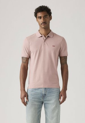 Mężczyzna ubrany w jasnoróżową koszulkę polo z krótkim rękawem i małym logo na piersi oraz jasnoniebieskie dżinsy na jednolitym tle.