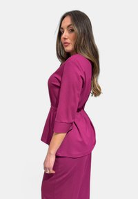 Top fucsia strutturato con maniche a tre quarti, vita cintrata e dettaglio plissettato sul retro. Tessuto morbido su uno sfondo semplice.