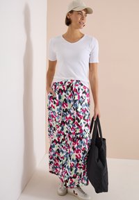 Femme portant un t-shirt blanc, une jupe longue à motifs multicolores, une casquette beige, des baskets blanches, tenant un grand sac cabas noir.