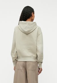 Φωτεινό μπεζ φούτερ με κουκούλα, κατασκευασμένο από απαλό υλικό, με χαλαρή εφαρμογή και ριμπ μανσέτες. Η οπίσθια όψη παρουσιάζει μαλακή υφή και σχεδιασμό.