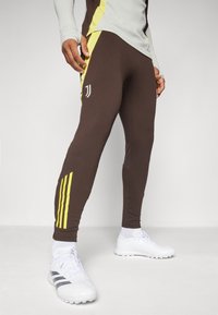 Un pantalon de sport marron avec des accents jaunes, une coupe slim et un logo blanc sur la cuisse. Porté avec des chaussures de sport blanches.