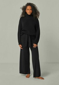 Schwarzer, gestrickter Rollkragenpullover kombiniert mit weit geschnittenen schwarzen Hosen, die einen stoffbezogenen Gürtel an der Taille haben. Weiche Textur und entspannter Schnitt.