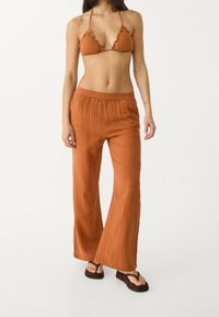 Bikini couleur rouille avec bords froncés et pantalon ample assorti. Tissu doux et texturé avec une ceinture élastique. Sandales plates.