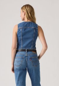 Veste sans manches en denim bleu moyen, dotée d'une coupe ajustée, de coutures visibles et d'une ceinture noire à la taille. Associée à un jean assorti.