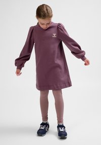 Lila Baumwollkleid mit Puffärmeln und rundem Halsausschnitt, verziert mit einem Logo auf der Brust, kombiniert mit hellen Leggings und Turnschuhen.