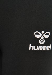 Schwarze sportliche Leggings aus dehnbarem Stoff mit einem weißen "hummel"-Logo und einem einzigartigen Muster in der Nähe des Logos.