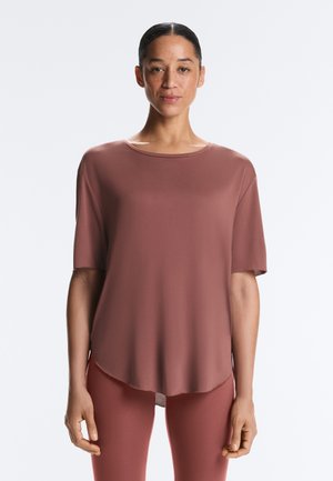 Žena s povučenom kosom, odjevena u labavu kratku majicu ljubičaste boje i hlače u istoj nijansi, stoji na jednostavnoj svijetloj pozadini.