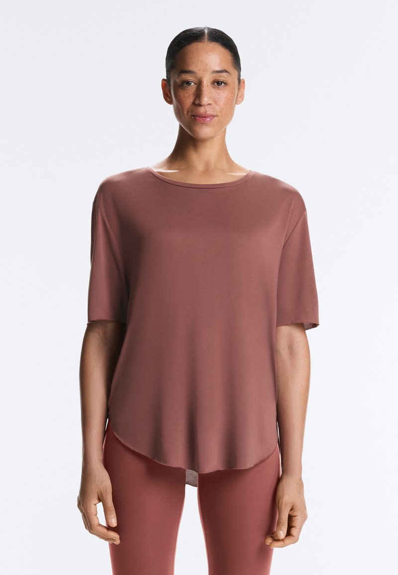 Femme aux cheveux attachés portant un haut mauve ample à manches courtes et un pantalon assorti, debout devant un fond clair uni.