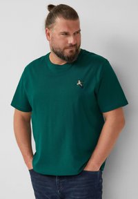 Beverde man met opgestoken haar, gekleed in een donkergroene T-shirt met een klein geborduurd vogeltje, met zijn handen in zijn zakken, kijkend opzij.