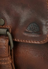 Dettaglio di una borsa in pelle marrone con superficie testurizzata, accessori in metallo e un logo di albero in rilievo sulla patta.