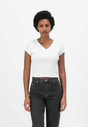 White ribbed crop top with a V-neck and short sleeves, paired with high-waisted dark jeans. Features a small embroidered logo on the side.