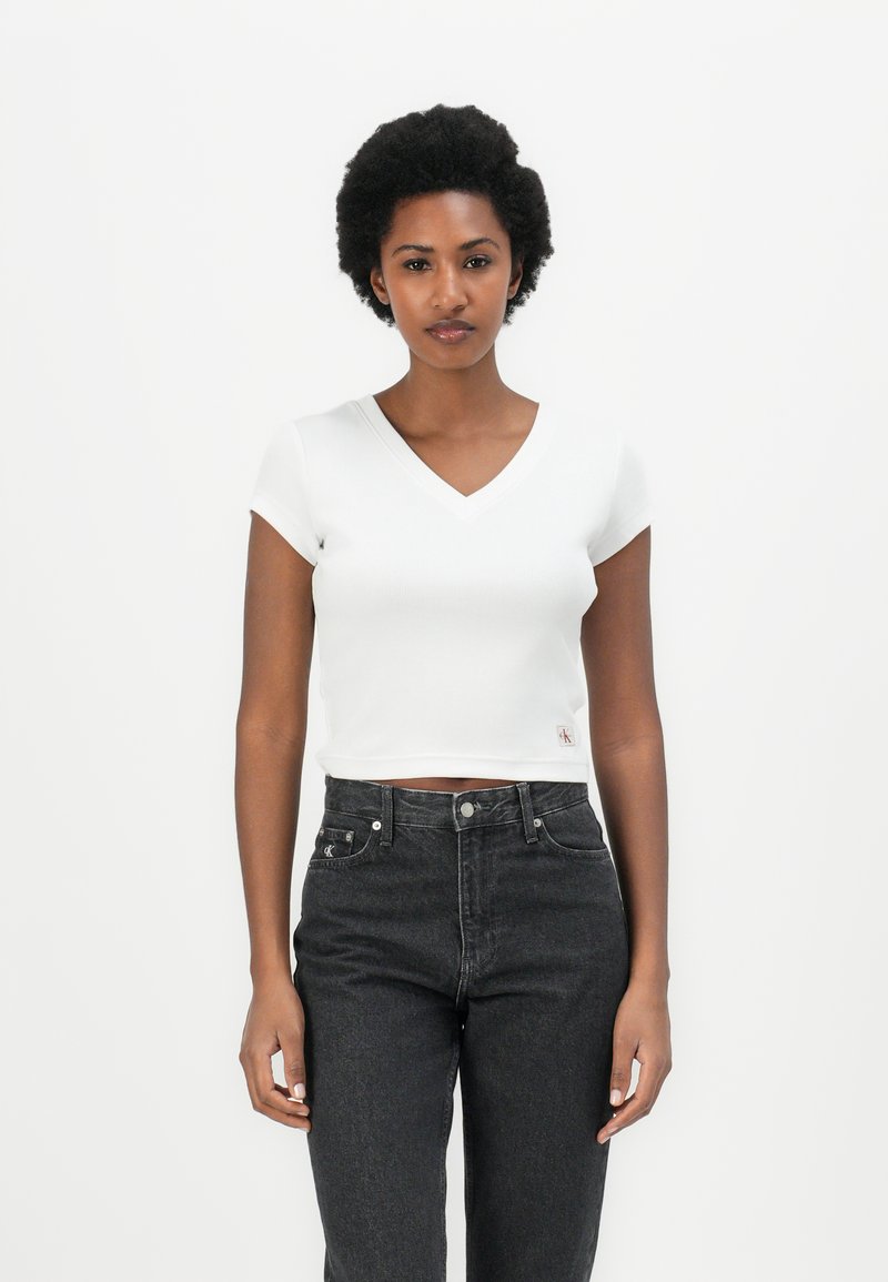 White ribbed crop top with a V-neck and short sleeves, paired with high-waisted dark jeans. Features a small embroidered logo on the side.
