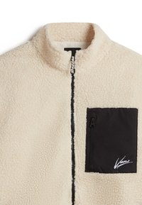 Fleecejacka i krämfärg med hög krage, svart dragkedja på bröstfickan och kontrasterande dragkedjefinish; mjuk textur och avslappnad design.