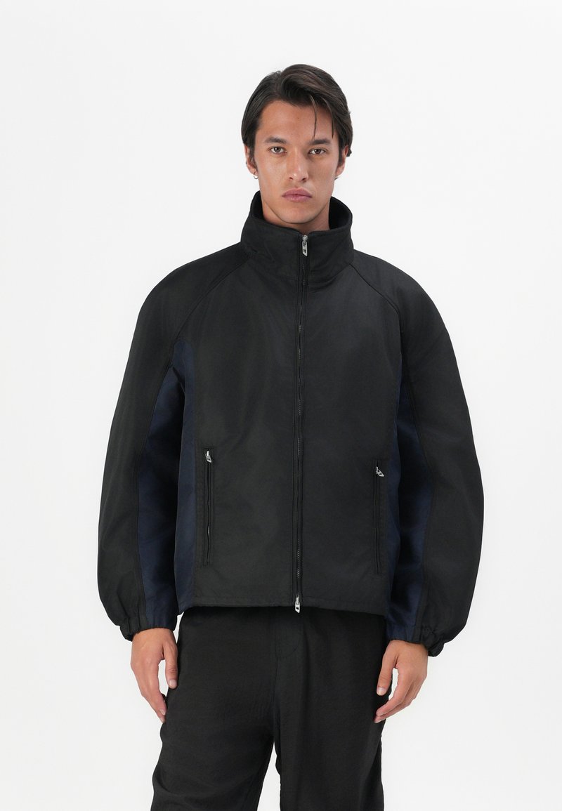 Black bomber jacket with a high collar, featuring a zip closure and side pockets; made of durable material with contrasting dark blue panels.