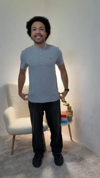 Uomo sorridente con capelli ricci che indossa una polo grigia e pantaloni neri in piedi in una stanza con una poltrona beige e un piccolo tavolo con libri e un vaso.