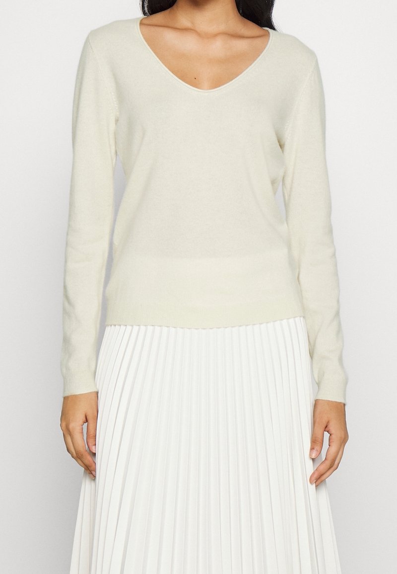 Femme portant un pull crème à col en V et manches longues, associé à une jupe midi plissée blanche, les mains reposant le long du corps.