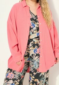 Light pink oversized button-up shirt made of textured fabric, worn over a black floral patterned outfit with vibrant colors and tropical designs.