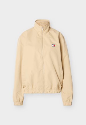 Beige jas met ritssluiting, hoge kraag, elastische manchetten en zoom, en een klein Tommy Jeans-logo op de linkerborst.