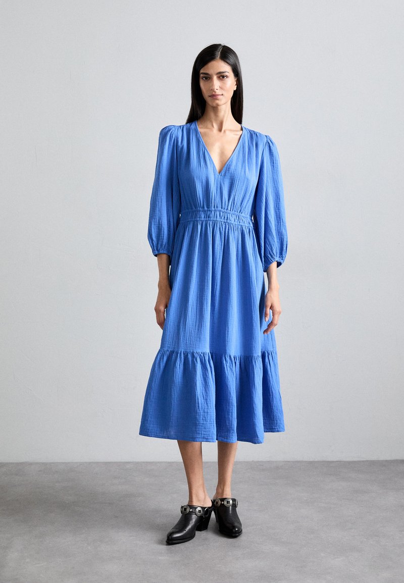 Blauwe, lange mouw, V-hals jurk met een gegeneraliseerde taille en een gelaagde rok. Geweven textuur, valt tot halverwege het scheenbeen. Gepaard met zwarte hoge klompen.