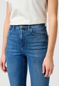 Jeans en denim bleu taille haute avec un design à cinq poches, bouton en laiton et léger effet vieilli sur le devant, avec une texture lisse.