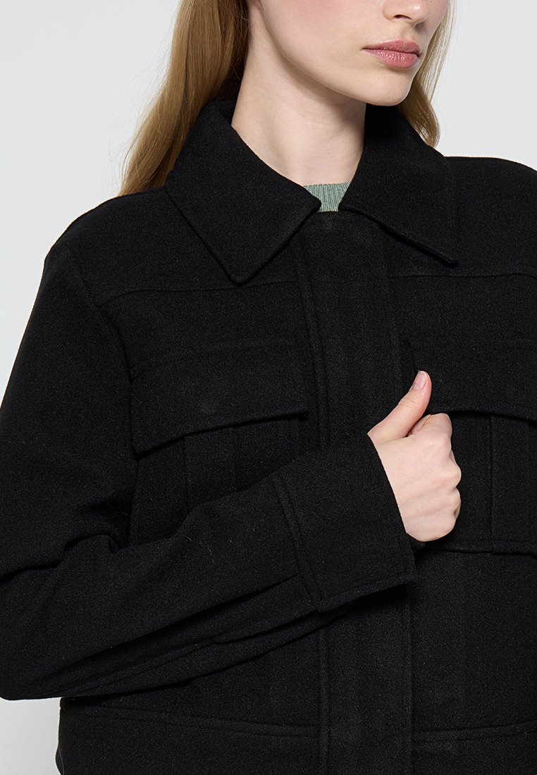 Veste en mélange de laine noire avec un col, des poches à l'avant et une finition texturée. Mains agrippant la veste près de la poitrine.