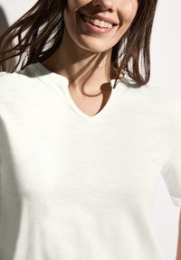T-shirt blanc à col en V avec une texture subtile, manches courtes et un petit détail de boutonnage près du cou. Tissu doux, coupe décontractée.