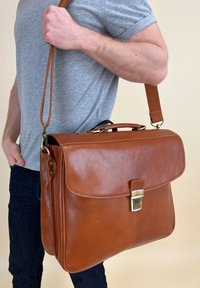 Mand bærer en grå t-shirt og en stor brun lædermapper med skulderrem og metalspænde.