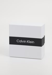 White rectangular box featuring a black band with the text "Calvin Klein" in white font. The design is minimalist and modern.