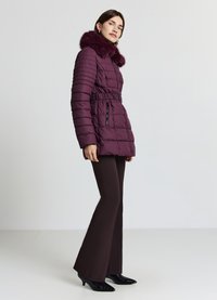 Veste matelassée bordeaux avec col en fausse fourrure, taille élastiquée et poches zippées, associée à un pantalon évasé marron et des talons noirs.