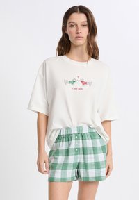 Donna che indossa una maglietta bianca larga con due dachshund vestiti in maglione e la scritta "Giornate cozy", abbinata a pantaloni corti a quadretti verdi e bianchi.