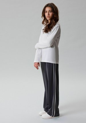 Joven mujer con cabello largo y castaño que lleva una camiseta blanca de manga larga, pantalones negros con rayas blancas y zapatillas blancas, de pie de lado.